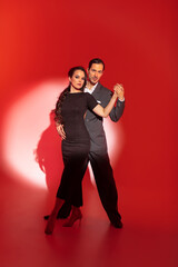 Creative portrait of tango dancers couple in dancing movement highlighted by a strip of light on red background. Handsome man in classic brown suit stands over a woman in elegant dress.
