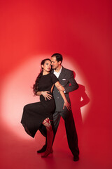 Creative portrait of tango dancers couple in dancing movement highlighted by a strip of light on red background. Handsome man in classic brown suit stands over a woman in elegant dress.