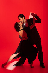 Creative portrait of tango dancers couple in dancing movement highlighted by a strip of light on red background. Handsome man in classic brown suit stands over a woman in elegant dress. - obrazy, fototapety, plakaty