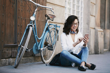 Young woman sitting on the ground using her mobile with her bicycle behind