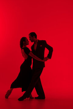 Dark silhouettes of tango dancers couple on red background. Man in classic suit and woman in elegant dress in dancing movement.