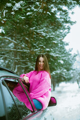 Fototapeta na wymiar Young cute girl in a pink sweater laughs and looks out of the car window on a winter road in the snow 