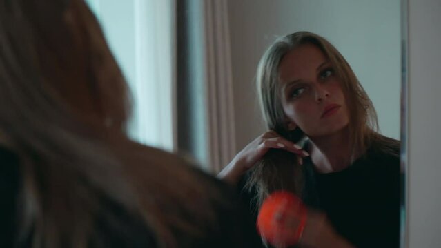Caucasian girl in a black dress combs her hair. A European woman with blond hair stands in front of a mirror with a comb. The lady is going on a date.