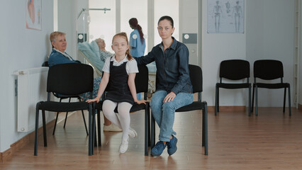 Portrait of mother and little girl in waiting room at clinic. Woman with child sitting in reception...