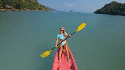 Pretty sportive blonde lady in stylish swimsuit sails on pink plastic canoe along azure ocean bay at exotic resort. Traveling to tropical countries. Girl is sailing on kayak in ocean, view from above.