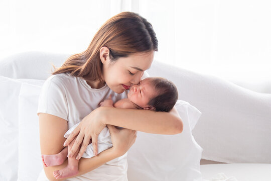 Selective focus of Beautiful Asian mother holding newborn baby in bed. Happy young woman hug and kissing adorable infant with love and tender. Mom spend time together with toddler at home.