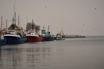Pier, four colorful ships after fishing are at the shore, seagulls are flying under them, the sea is calm, the sky is grey