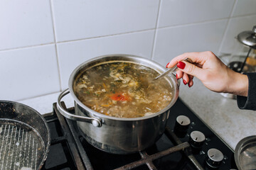 The girl, the cook, prepares soup from vegetables in boiling water, pouring green dill from a spoon into a saucepan. Food preparation.