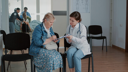 Aged patient signing checkup papers on clipboard for doctor in waiting area. Elder woman with sickness having medical appointment and consultation with physician in waiting area.