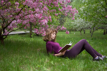 young woman, student, reads book with interest, lying under tree on spring day in park. girls -...