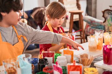 Boy pointing with his finger at the glass with paints while drawing near his brother or friend