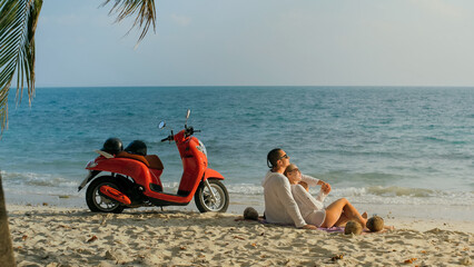 Scooter road trip. Lovely couple on red motorbike in white clothes on sand beach. Just married people kiss hugs walking near the tropical palm trees, sea. Wedding honeymoon by ocean. Motorcycle rent.