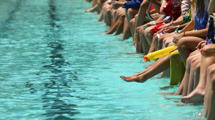 A bright aqua blue swimming pool with students sitting dangling their feet and toes in the water...