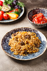 Pilaf with lamb meat in large dish, plate of tomato salad and dish with vegetables and greens