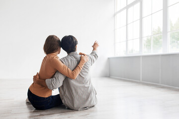 Glad happy dreaming young european couple hugs, planning future interior show at window sit on floor