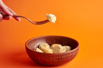A woman's hand holds a dumpling on a fork in front of a bowl. Orange background