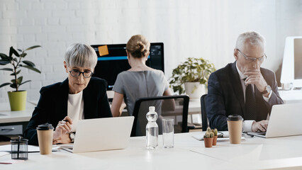 Mature business people using laptops near drinks in office