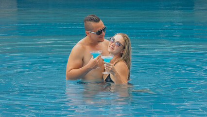 Fototapeta na wymiar The loving couple hugs and kisses, drinking blue cocktail alcohol liquor in swimming pool at hotel outdoor. Portrait of caucasian man and woman. Creative hairstyles bodybuilder, swimsuit, sunglasses.