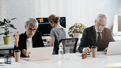 Mature businesswoman looking at laptop near drinks and colleague in office