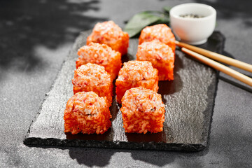 Baked Maki sushi on dark slate. Hot california maki with crab. Sushi roll with masago outside, baked cheese and crab topped. Style concept japanese menu with black background, leaves and hard shadow.
