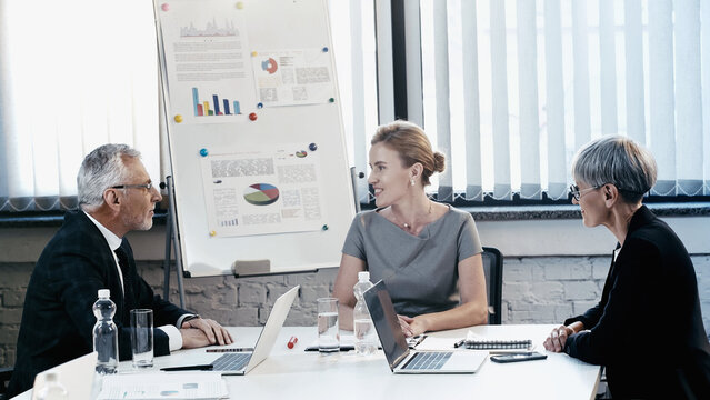 Smiling businesswoman talking to mature colleagues near papers and devices in office
