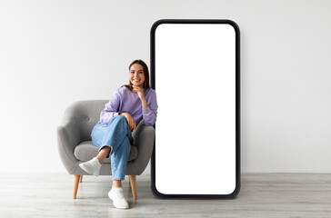 Happy woman sitting on chair by big smartphone with empty screen, recommending online services, app...