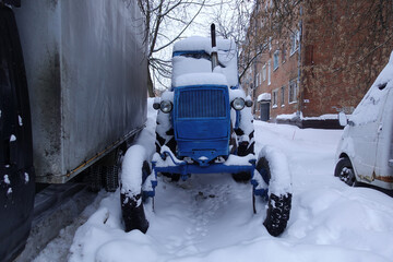 An old Soviet tractor in the snow among the cars in the parking lot.