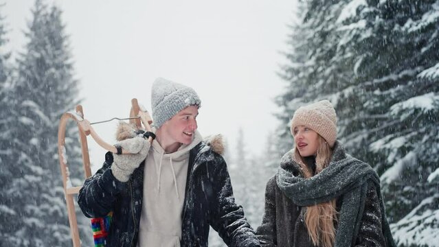 Happy young couple in love walking and carrying sledge in wintery nature under falling snow.