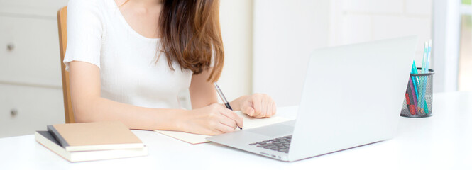 Young asian woman writing on notebook for planning working and using laptop computer on desk at...