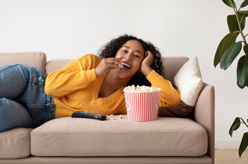 Young black woman relaxing on couch, watching TV and eating popcorn at home
