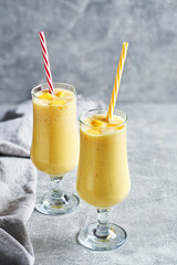 Mango banana yellow smoothie healthy breakfast in drinking glass. Detox yellow smoothie made with fresh banana and mango in two drinking glass with straws on gray background. Simple yellow smoothie.