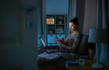 education, technology and people concept - teenage student girl with notebook and laptop computer learning in bed at home at night