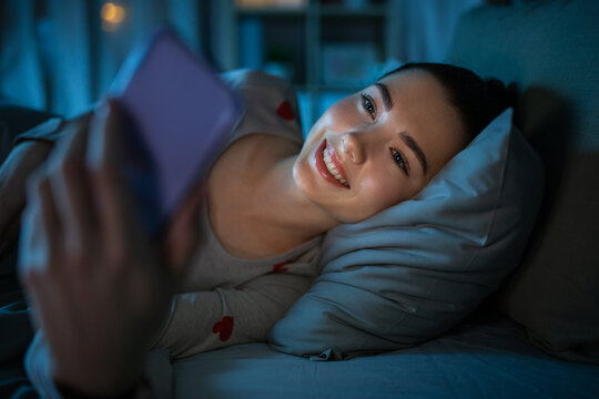 technology, bedtime and rest concept - smiling teenage girl in pajamas with smartphone lying in bed at night