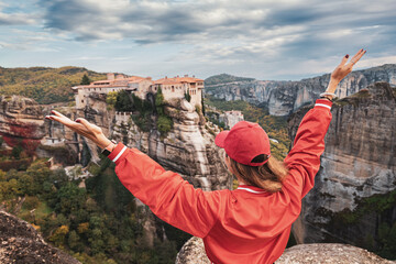 Happy traveler girl with raised arms standing on top of a cliff and admires view of the famous...