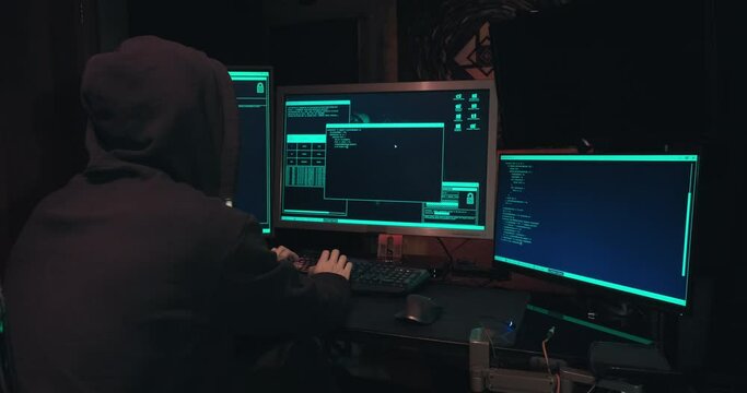 Hacker in a hood sits in front of computer screens and hacks databases. The concept of cyberterrorism, server hacking and cyberattacks. The room is dark, a person is typing code into the command line