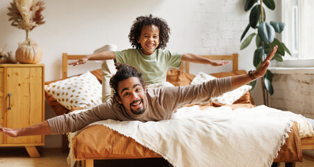 Happy african american family father and son in flying pose lying on bed at home