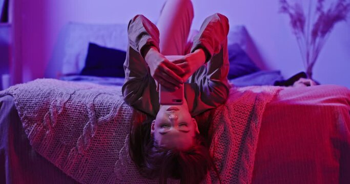 Teenage girl relaxing on comfortable bed and messaging on social media via mobile phone in room with pink neon light