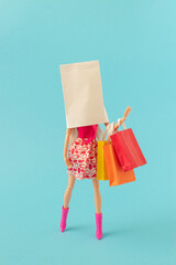 Creative look of stylish girl with white paper bag over head holding colorful bags on pastel blue background. Minmal creative trandy young woman concept. Copy space.