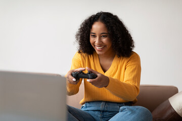 Cool young black woman with gaming joystick playing computer game on laptop pc at home