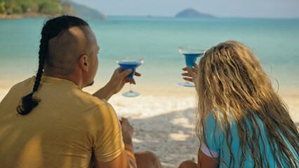 The love couple is holding a glass of blue curacao cocktail, on sea. Man with plait and blonde...