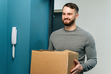 Portrait of friendly bearded courier male holding cardboard box parcel in hands standing on doorway...