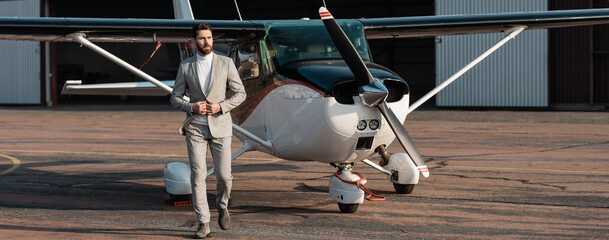 full length of bearded businessman in suit walking near modern helicopter outdoors, banner.