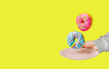 Fototapeta na wymiar Two multi-colored donuts levitate above a plate held by a hand on a yellow background. The concept of unhealthy eating