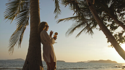 SILHOUETTE DARK GOLD SUNSET VIEW: Pretty woman drinking pineapple cocktail Pina Colada near palm...