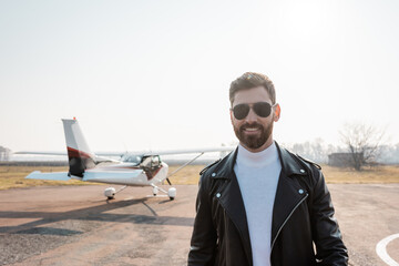 cheerful pilot in black leather jacket and sunglasses near helicopter.