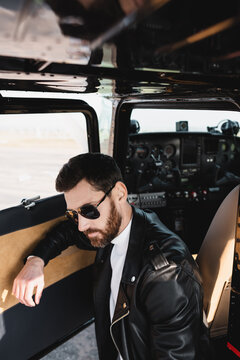 bearded pilot in stylish sunglasses and black leather jacket near opened door of helicopter.