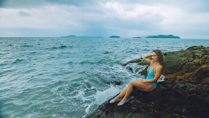 Alone attractive woman walk near a rock reef hill in stormy morning rain cloudy sea. Girl in turquoise swimsuit. Concept resort outdoor relax, vacation, loneliness. Dark dramatic view