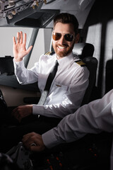 cheerful pilot in sunglasses waving hand near co-pilot in airplane.