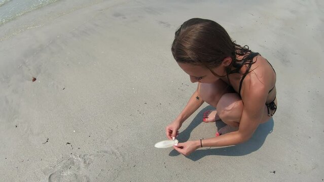 A tanned caucasian girl sits on a white sandy beach. A European woman in a bathing suit found a shell on the seashore.