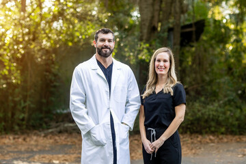 A doctor with dark hair and a beard in black scrubs and a white lab coat standing outside in a natural green environment talking to another medical professional